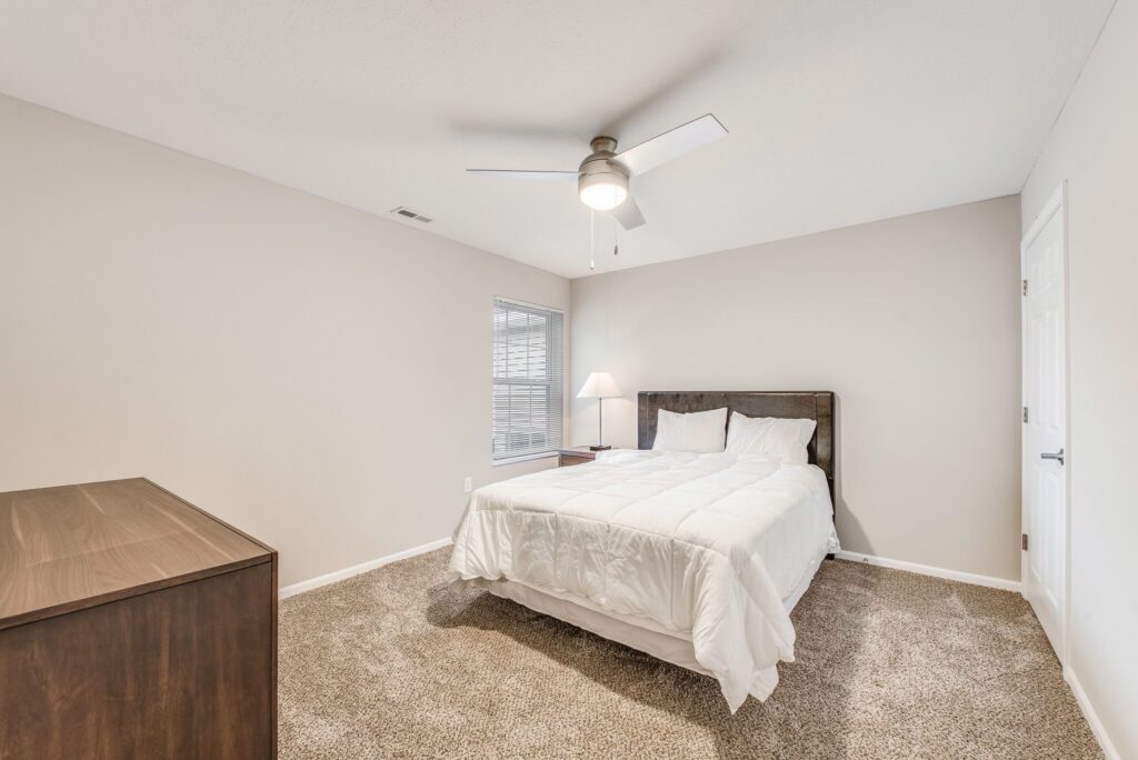 Bedroom with beige carpet, white walls, a ceiling fan, a double bed with white bedding, a wooden dresser, and a window with blinds letting in natural light.