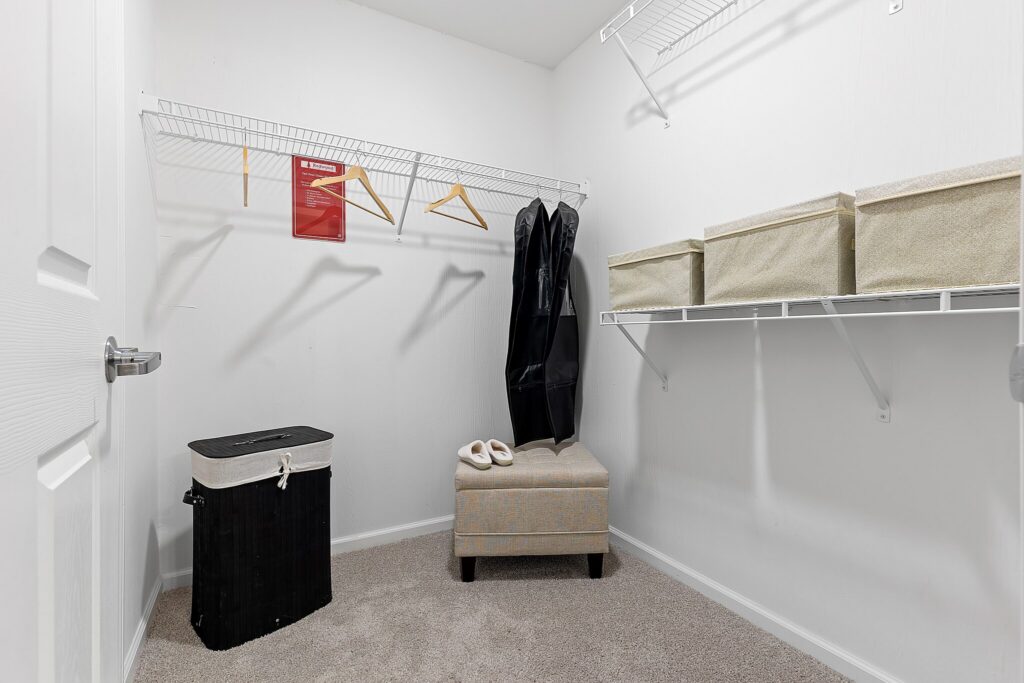 Walk-in closet with beige carpet, white walls, wire shelves with storage bins, a laundry hamper, ottoman, two garment bags, slippers, and wooden hangers.