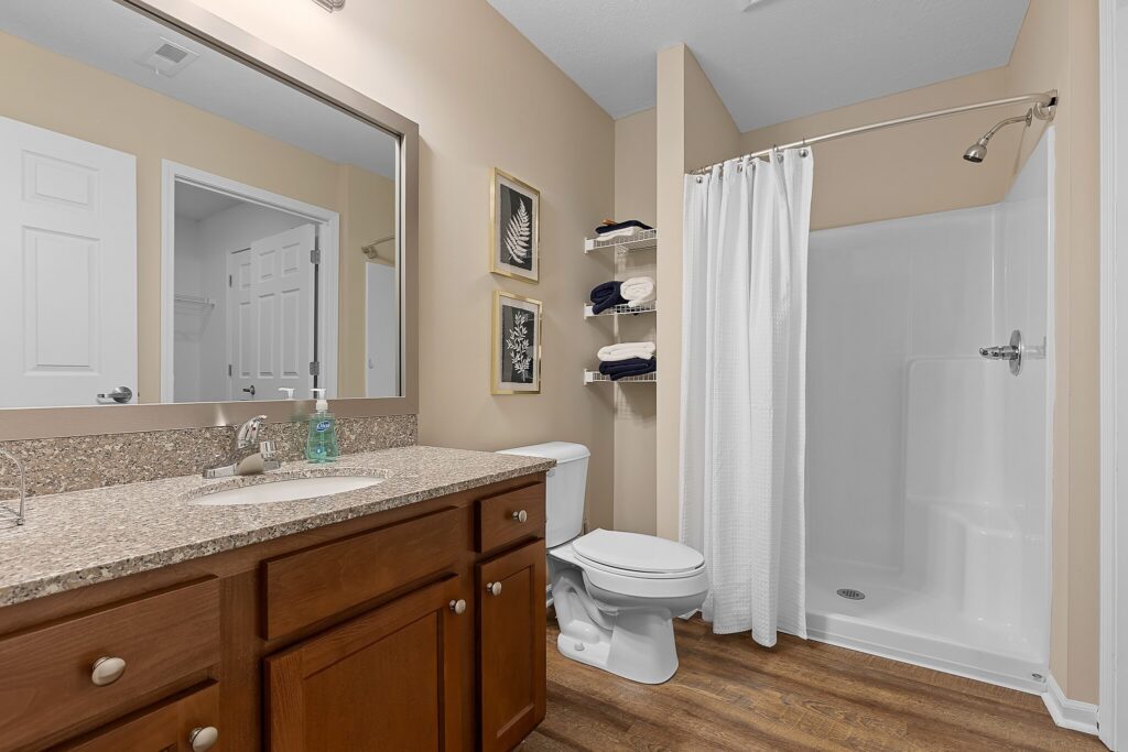 Modern bathroom with a wooden vanity, granite countertop, sink, toilet, open shelving with towels, and a walk-in shower with a white curtain.