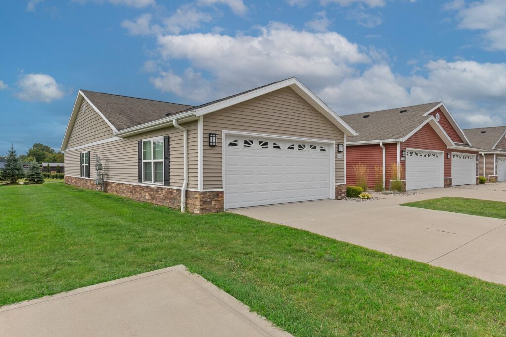 Single-story house with beige siding, stone trim, and a white double garage door, located in a suburban neighborhood with a green lawn and driveway.