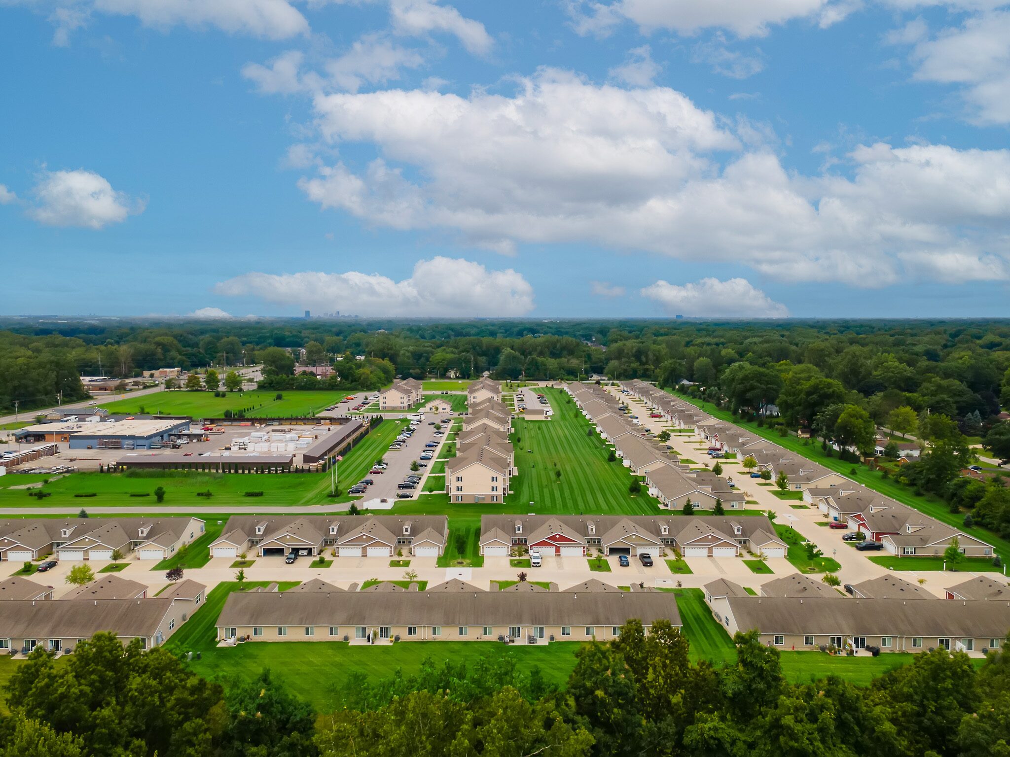 Aerial view of a suburban residential area with rows of houses, well-maintained lawns, and parking areas under a partly cloudy sky.