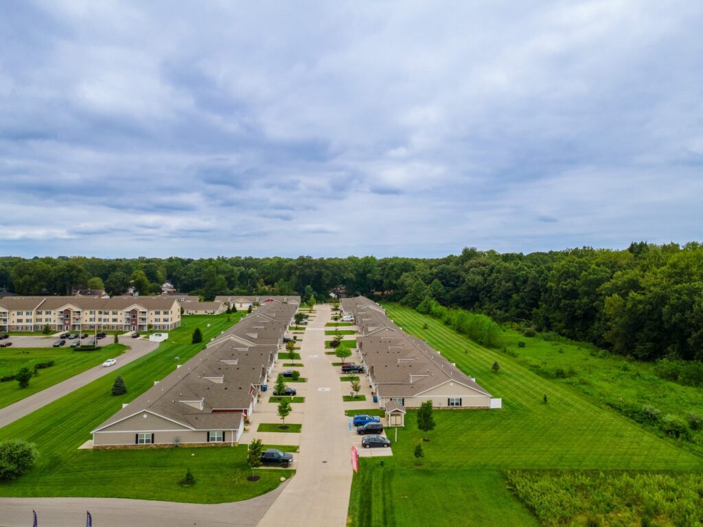 Aerial view of a residential neighborhood with rows of single-story homes, parked cars, green lawns, and surrounding trees under a cloudy sky.
