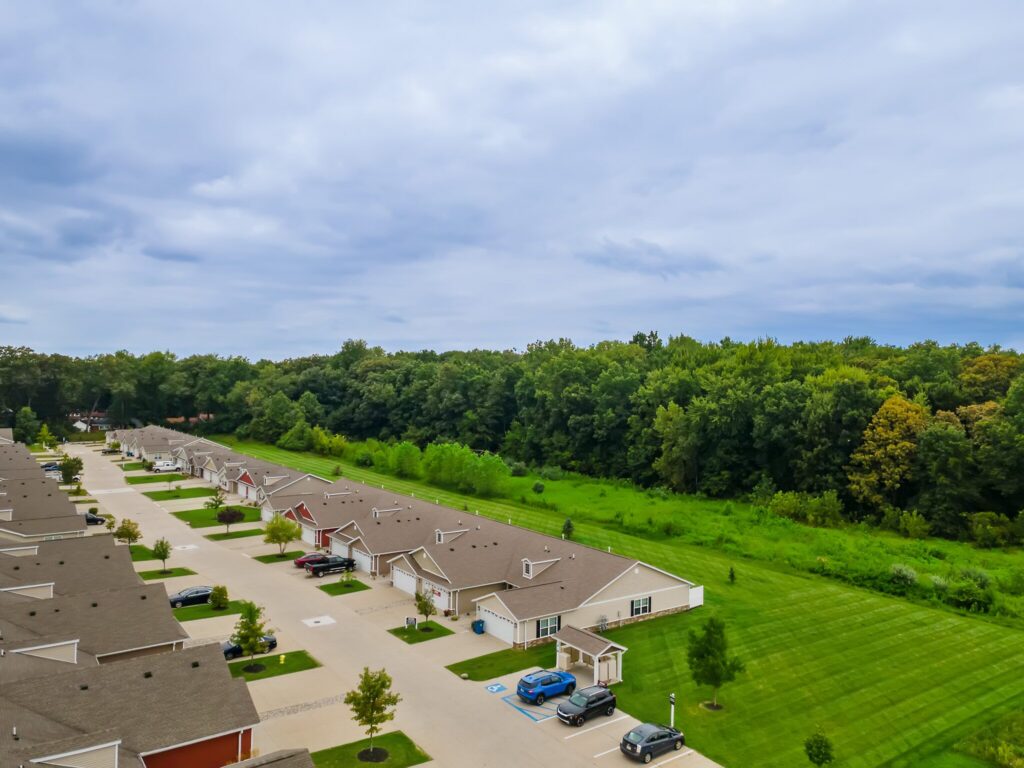 A row of single-story houses with driveways faces a grassy area and dense trees under a partly cloudy sky. Several cars are parked in the driveways.