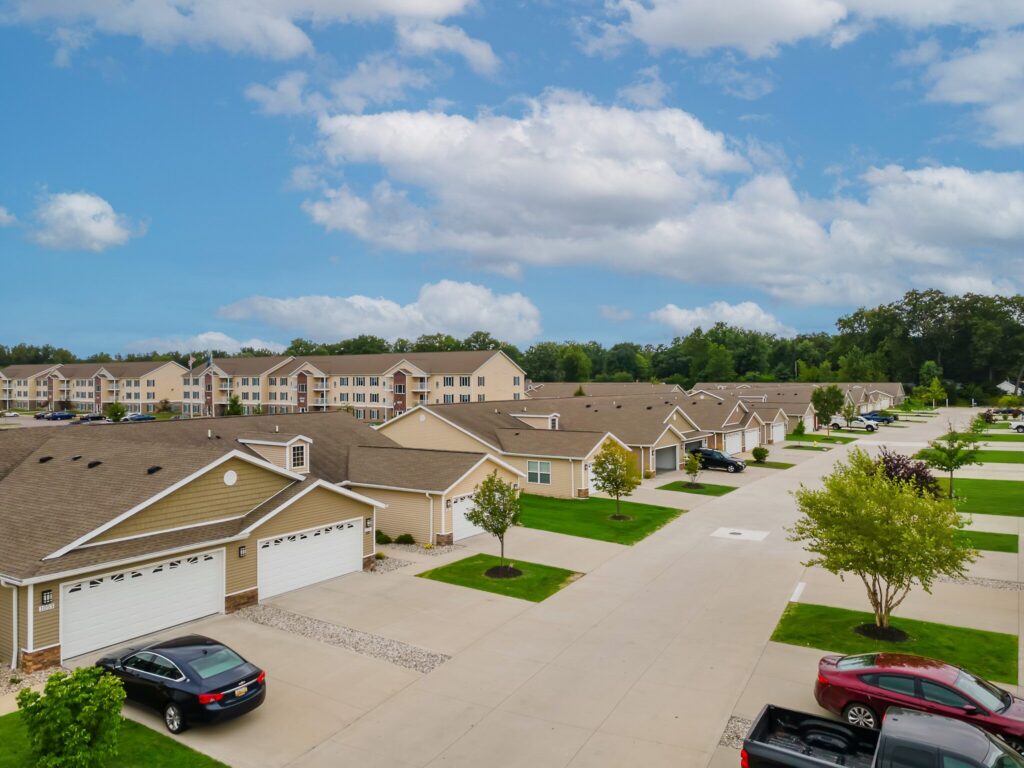 A suburban neighborhood with single-story houses, driveways, parked cars, green lawns, and trees lining a wide street under a partly cloudy sky.