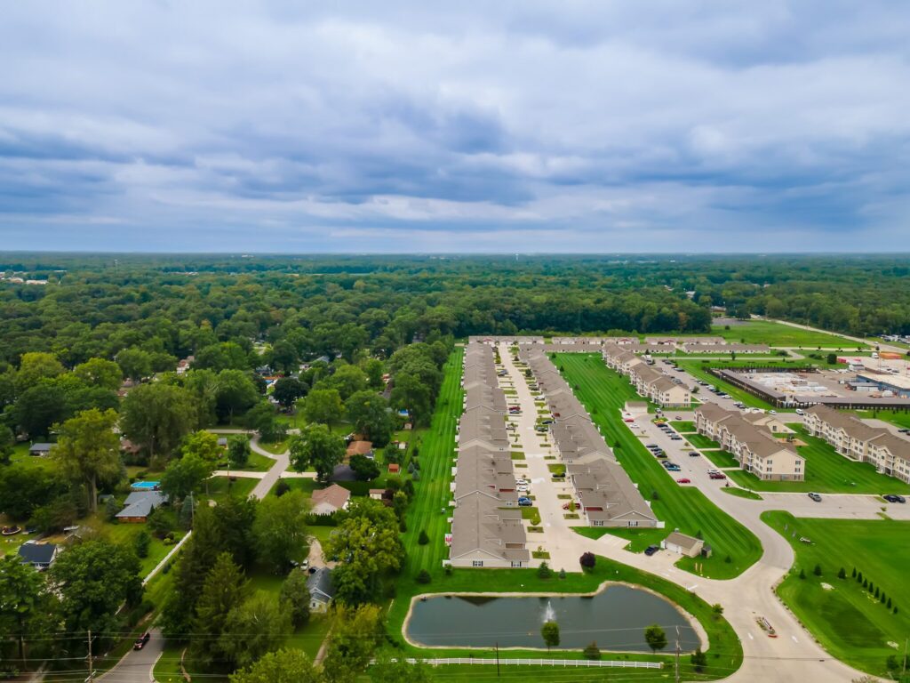 Aerial view of a suburban neighborhood with rows of houses, green lawns, roads, a small pond, and dense trees in the background under a cloudy sky.