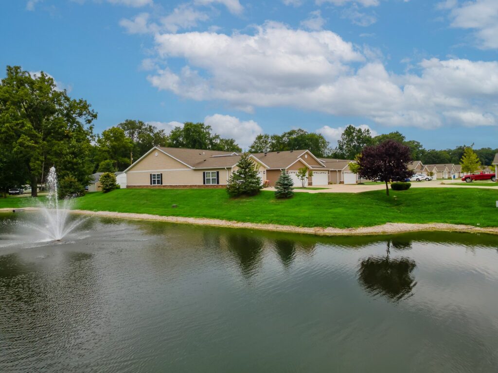 Single-story residential buildings with garages are seen across a pond with a water fountain, surrounded by trees and greenery under a partly cloudy sky.