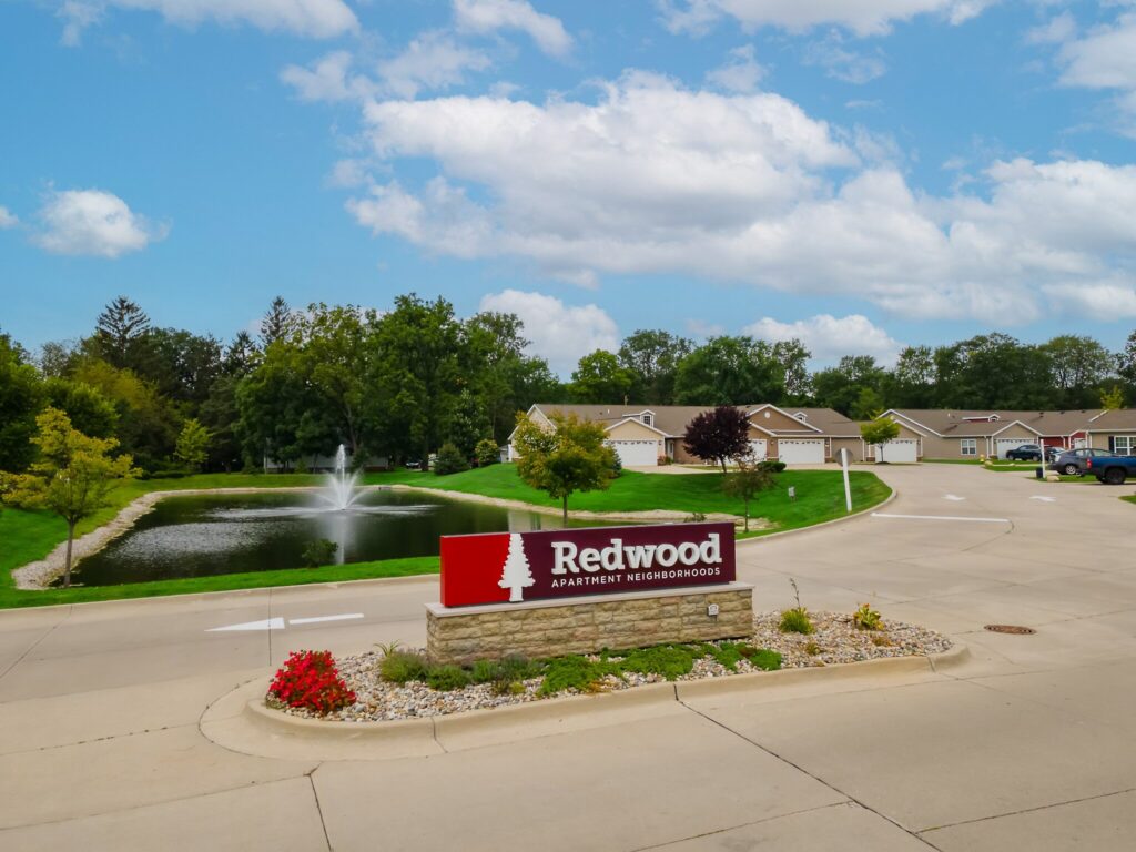 Redwood Apartment Neighborhoods sign in front of a pond with a fountain, surrounded by single-story residential buildings and trees under a partly cloudy sky.