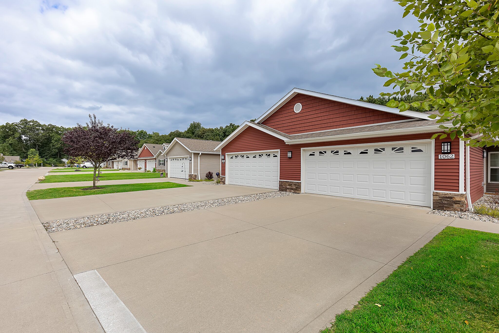 Row of single-story suburban houses with attached garages and well-maintained lawns under a cloudy sky. Wide, clean driveways line the street.