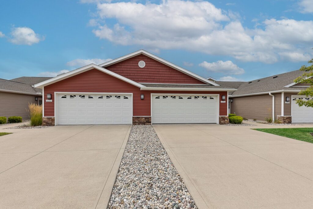 A duplex with red and tan siding, two white double garages, and a stone path dividing the driveway, set under a partly cloudy sky.