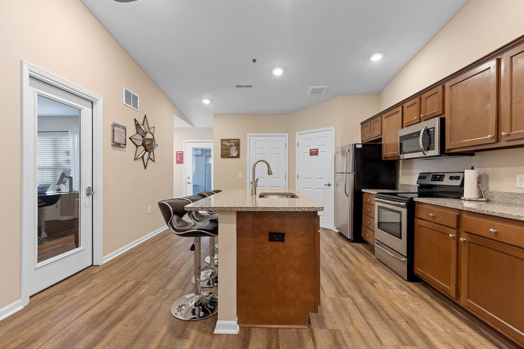 Modern kitchen with brown cabinets, granite countertops, stainless steel appliances, island with sink, and two bar stools; adjacent room visible through glass door.