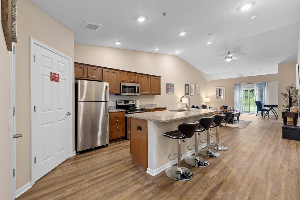 Modern open-concept kitchen and living area with stainless steel appliances, granite countertop island, three barstools, wood flooring, and sliding glass doors leading to a patio.