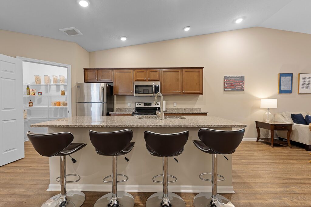 Modern kitchen with brown cabinets, stainless steel appliances, a large island with four bar stools, and open living area with a sofa and wall art in the background.