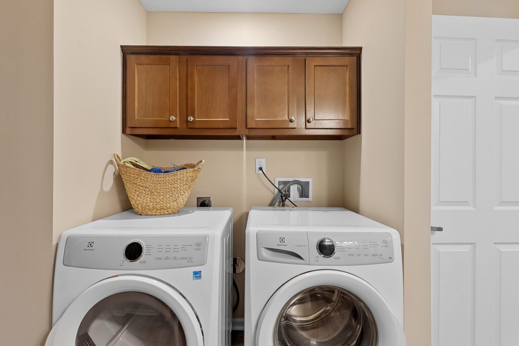 A laundry room with a front-loading washer and dryer, a wicker basket on top, and wooden cabinets mounted above against a beige wall.