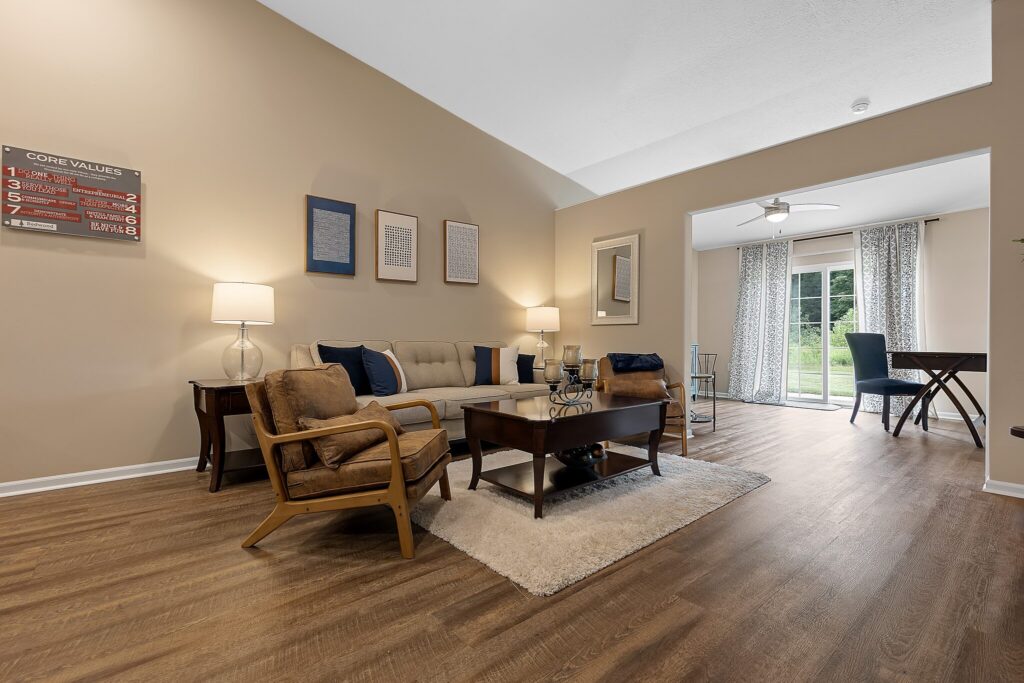 Spacious living room with beige walls, wood flooring, a sofa, armchairs, coffee table, lamps, and wall art, leading to a dining area with glass doors opening to a backyard.