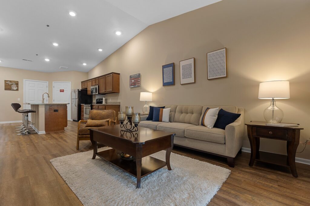 Modern open-concept living room and kitchen with wooden flooring, neutral walls, a beige sofa, coffee table, bar stools, and overhead lighting.