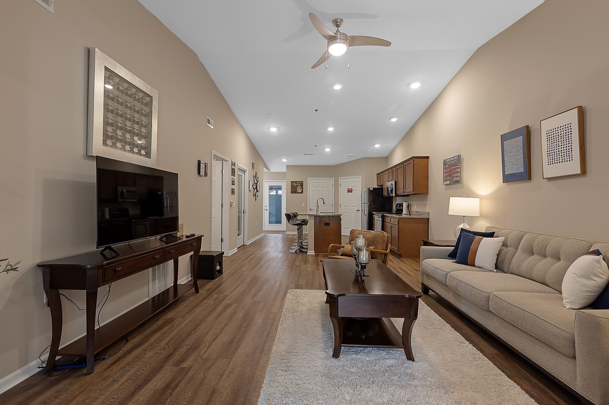 Open-concept living room and kitchen with wood flooring, beige walls, modern furniture, a wall-mounted TV, and recessed ceiling lights.