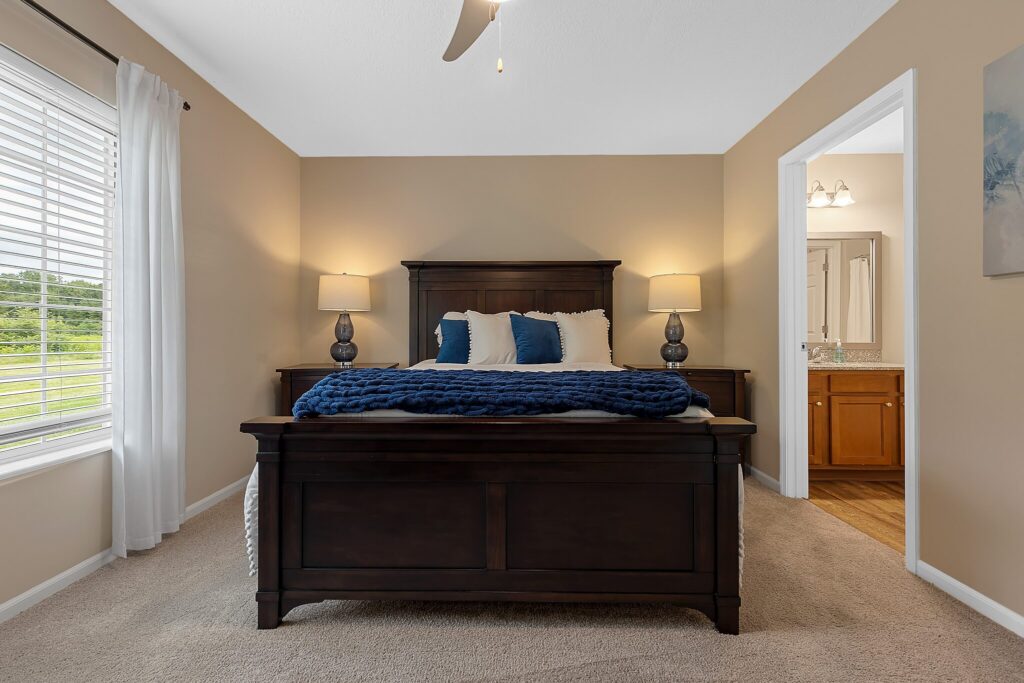 A bedroom with a dark wooden bed, blue and white bedding, two bedside tables with lamps, beige walls, carpeted floor, window with white curtains, and a bathroom visible.