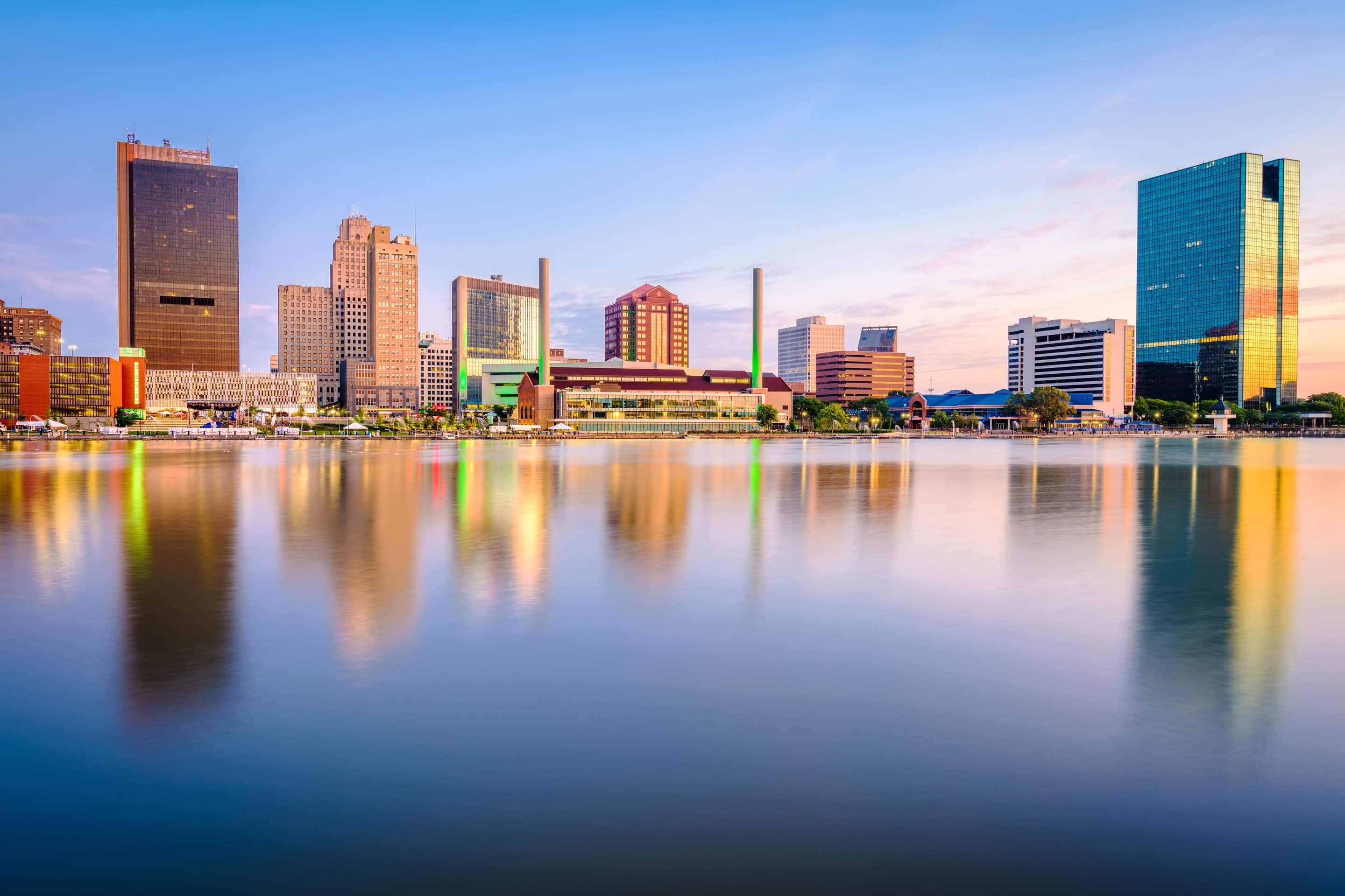 Downtown city skyline with modern and historic buildings reflected in a calm river under a clear sky at sunset.