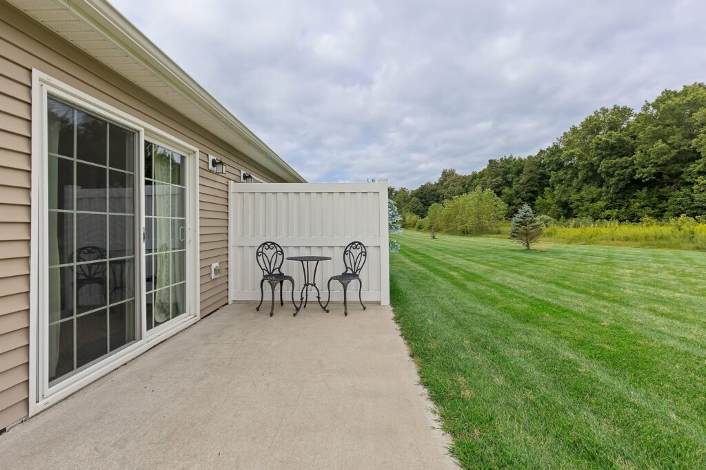 A small patio with a black metal table and two chairs sits outside a sliding glass door, next to a white privacy fence and overlooking a large grassy yard with trees.