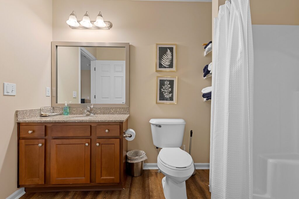 Bathroom with a wooden vanity, granite countertop, sink, toilet, trash can, towel shelves, framed leaf art, and a white shower curtain.