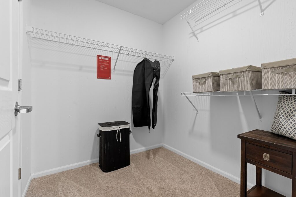 A walk-in closet with wire shelving, a closed hamper, three storage boxes, a hanging garment bag, and a wooden table with a patterned pillow on top.