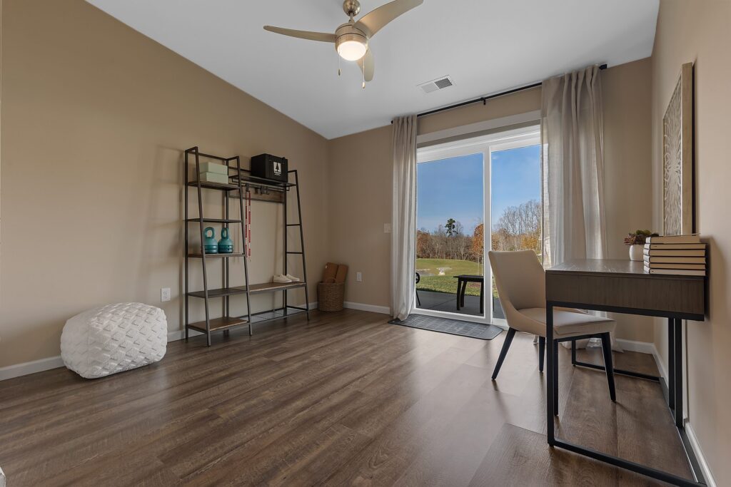A home office with a desk and chair near a window, open shelving against a beige wall, wood flooring, and glass doors leading to a backyard.