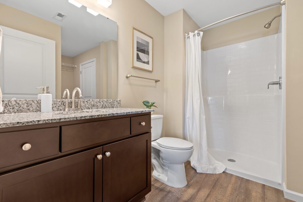 Modern bathroom with a granite countertop vanity, toilet, wall art, towel rack, and a walk-in shower with a white curtain; wood-style flooring throughout.