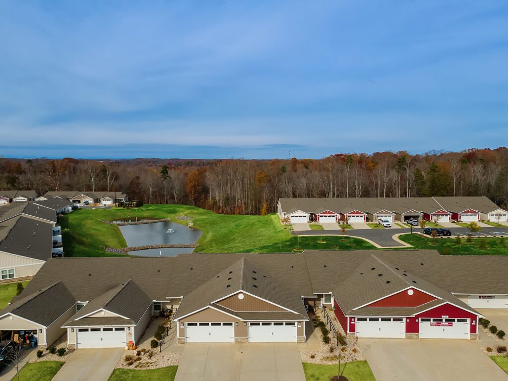 Aerial view of suburban single-story homes with driveways, a small pond, green lawns, and a wooded area in the background.