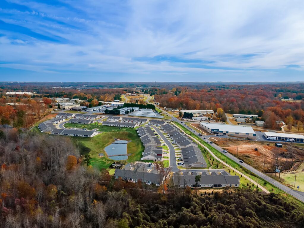 Aerial view of a suburban neighborhood with rows of houses, surrounding roads, trees with autumn foliage, and commercial buildings in the background under a partly cloudy sky.