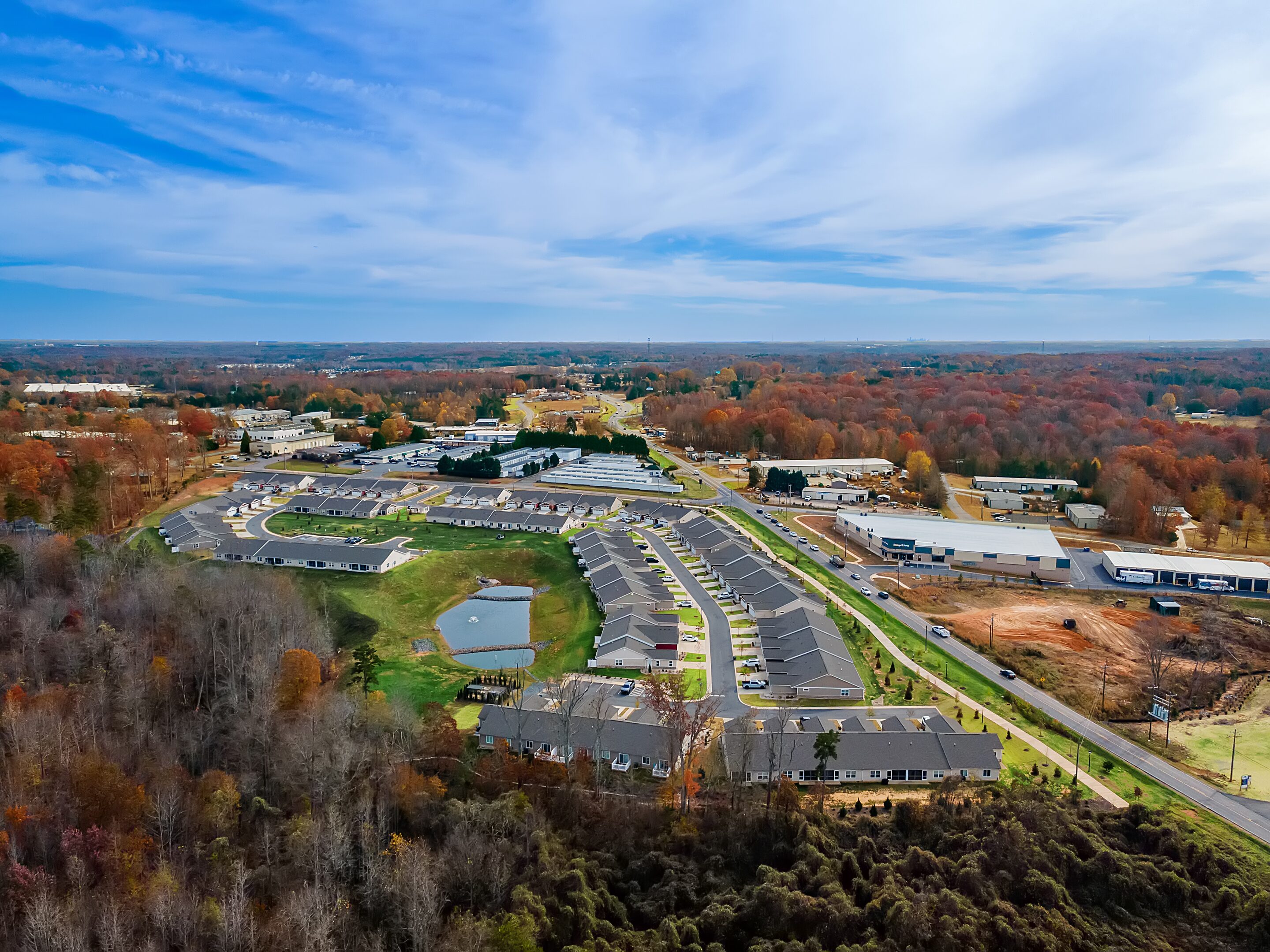 Aerial view of a suburban neighborhood with rows of houses, surrounding roads, trees with autumn foliage, and commercial buildings in the background under a partly cloudy sky.