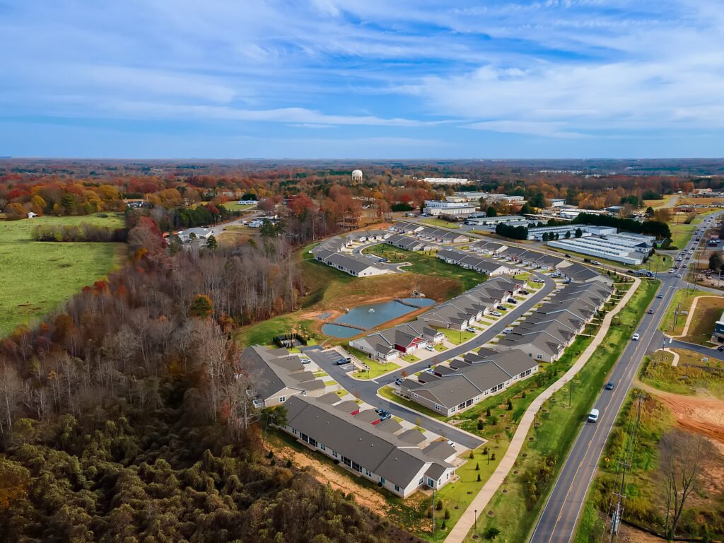 Aerial view of a suburban neighborhood with rows of houses, a pond, roads, and surrounding trees under a partly cloudy sky.