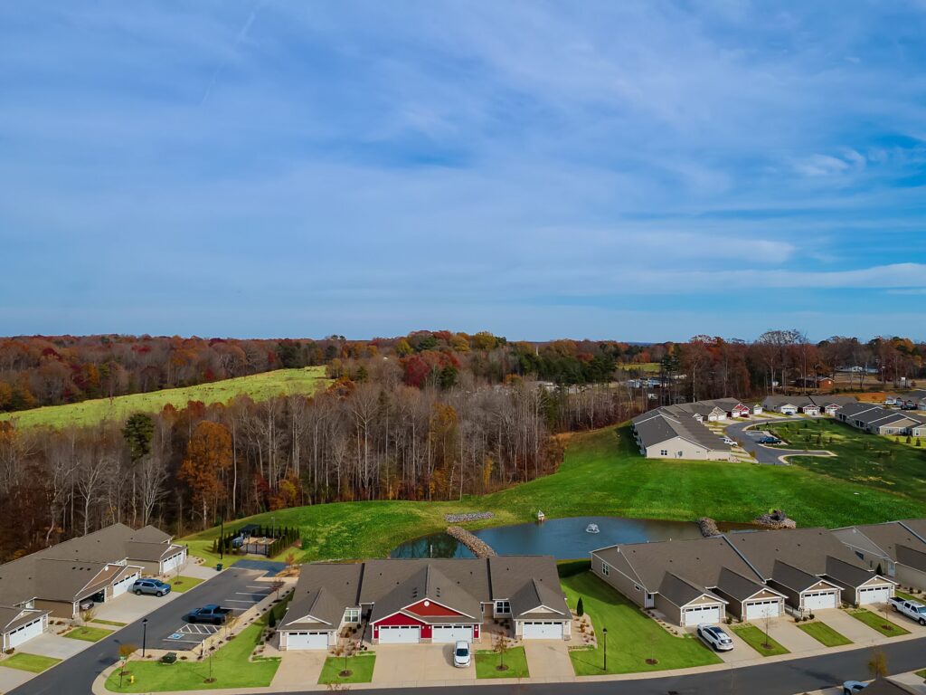 Aerial view of a suburban neighborhood with rows of houses, green lawns, a small pond, and a forested area in the background under a blue sky.
