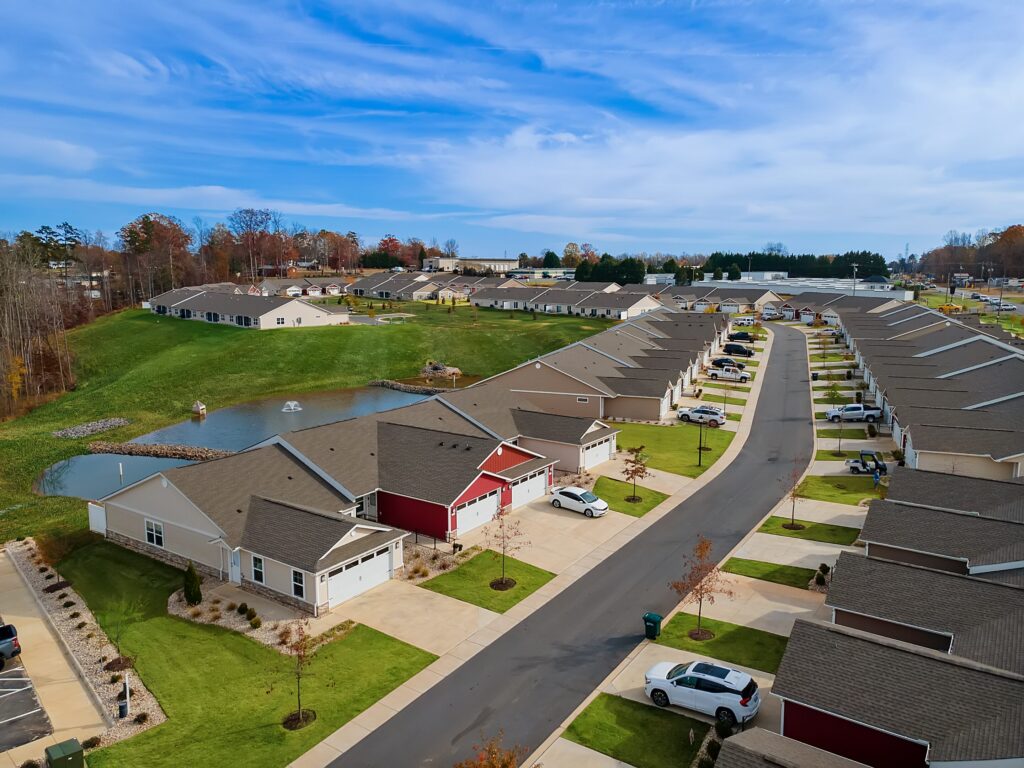 Aerial view of a suburban neighborhood with rows of single-story houses, driveways, parked cars, and a small pond with a fountain.
