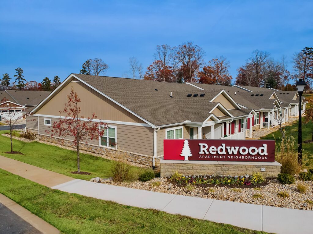 Single-story apartment buildings with tan siding and a sign in front that reads "Redwood Apartment Neighborhoods," surrounded by grass and landscaping.