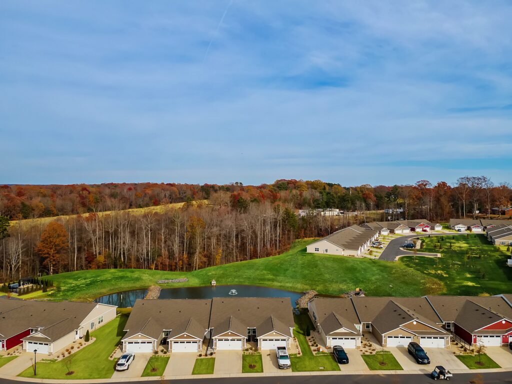 Aerial view of a suburban neighborhood with rows of houses, a pond, green lawns, and a wooded area with autumn foliage in the background under a blue sky.