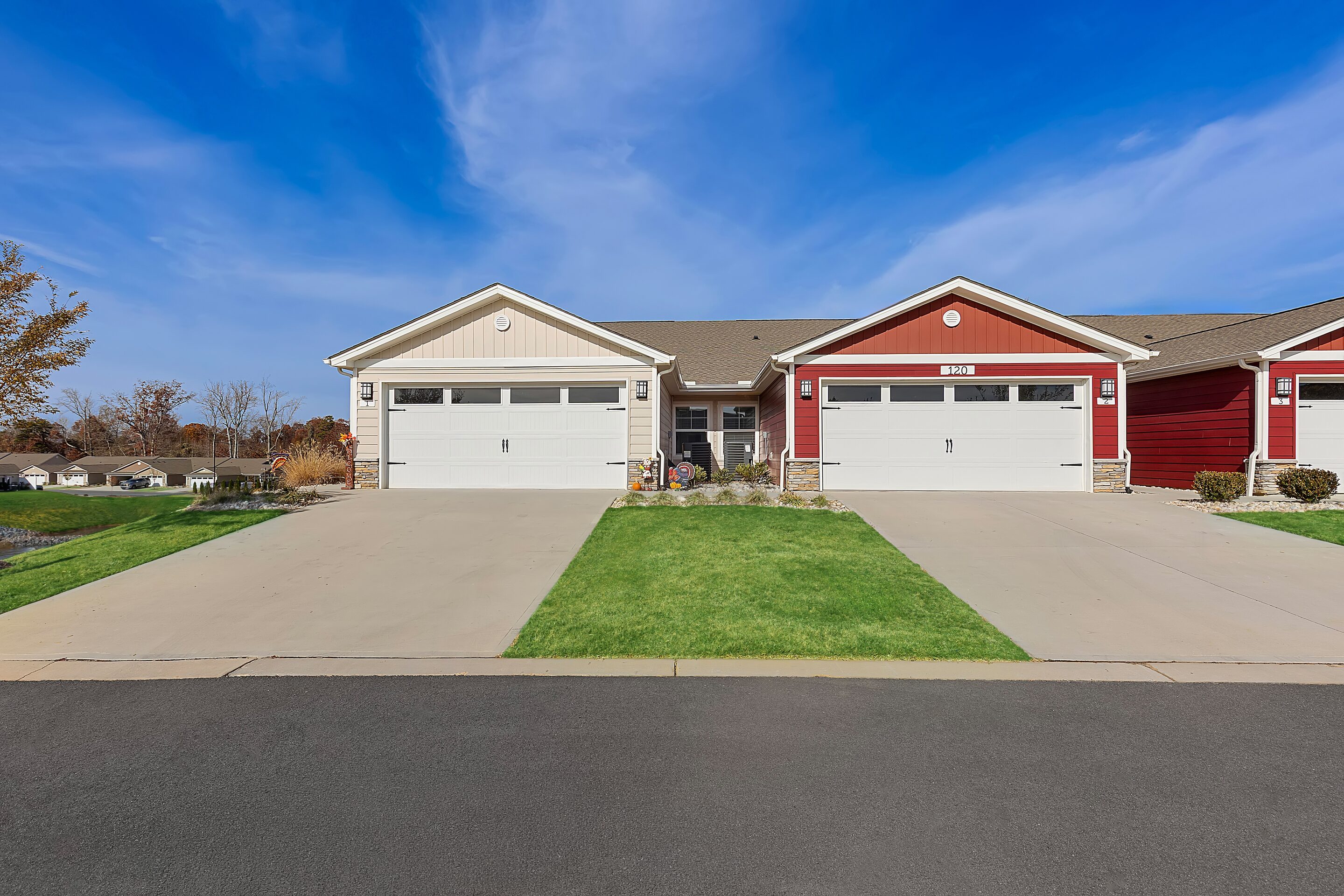 Two attached single-story houses with double garages, beige and red exteriors, green lawns, and a clear blue sky above.
