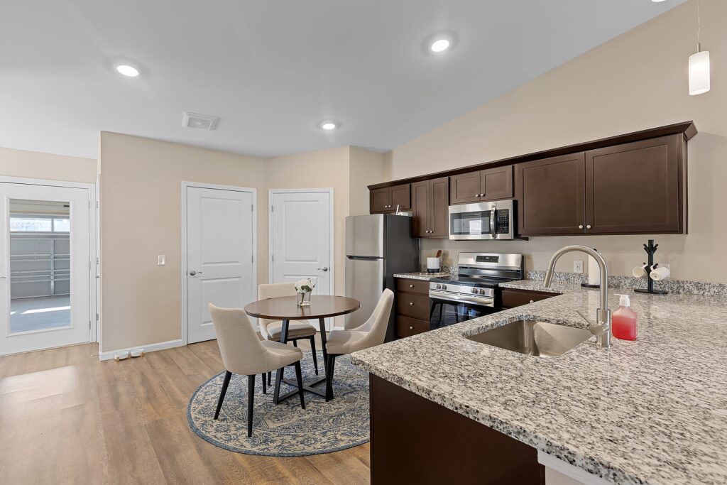 Modern kitchen and dining area with granite countertops, stainless steel appliances, dark wood cabinets, and a round dining table with four chairs on a blue rug.