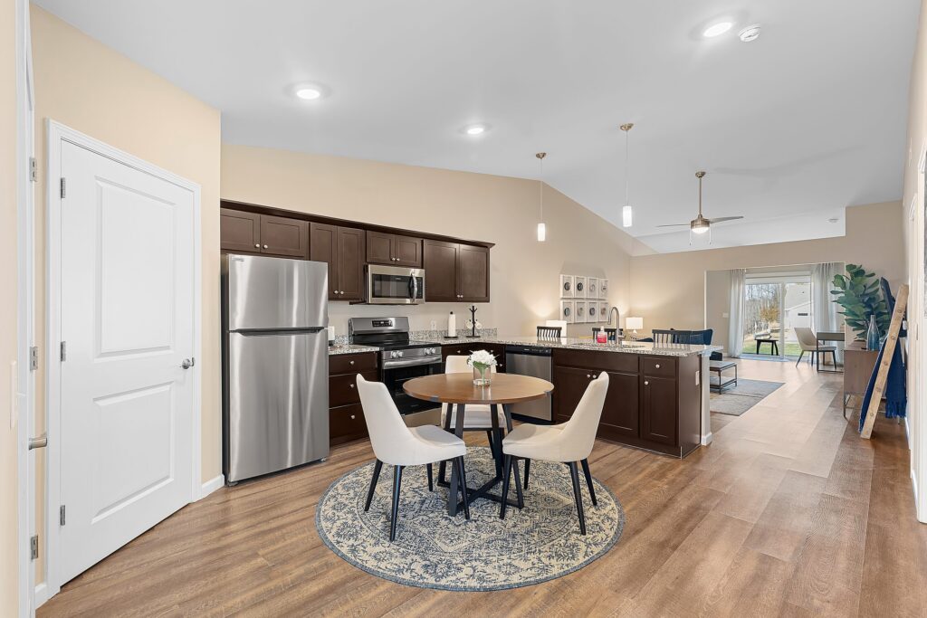 Modern open-concept kitchen and dining area with dark wood cabinets, stainless steel appliances, round table with two chairs, and view into living room with large windows.