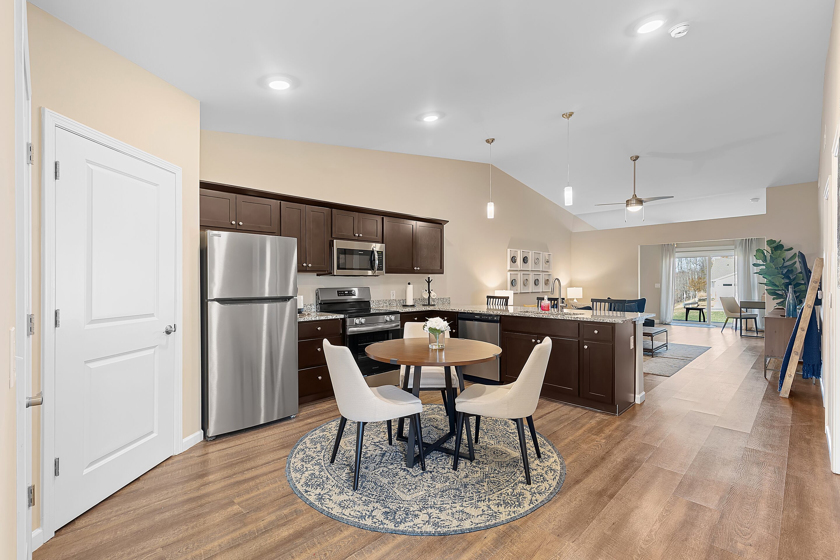 Modern open-concept kitchen and dining area with dark wood cabinets, stainless steel appliances, round table with two chairs, and view into living room with large windows.