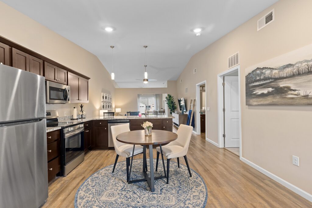 Modern kitchen and dining area with stainless steel appliances, dark wood cabinets, a round table on a rug, and wall art; light wood flooring throughout the space.
