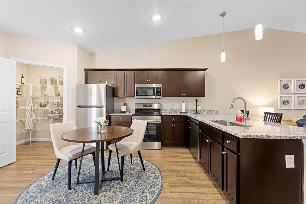 Modern kitchen with dark wood cabinets, stainless steel appliances, a round dining table with two chairs, granite countertops, and an open pantry with shelves.