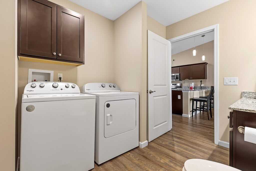 Laundry room with a washing machine and dryer, brown cabinets, beige walls, wood flooring, and a view into a kitchen with stainless steel appliances.