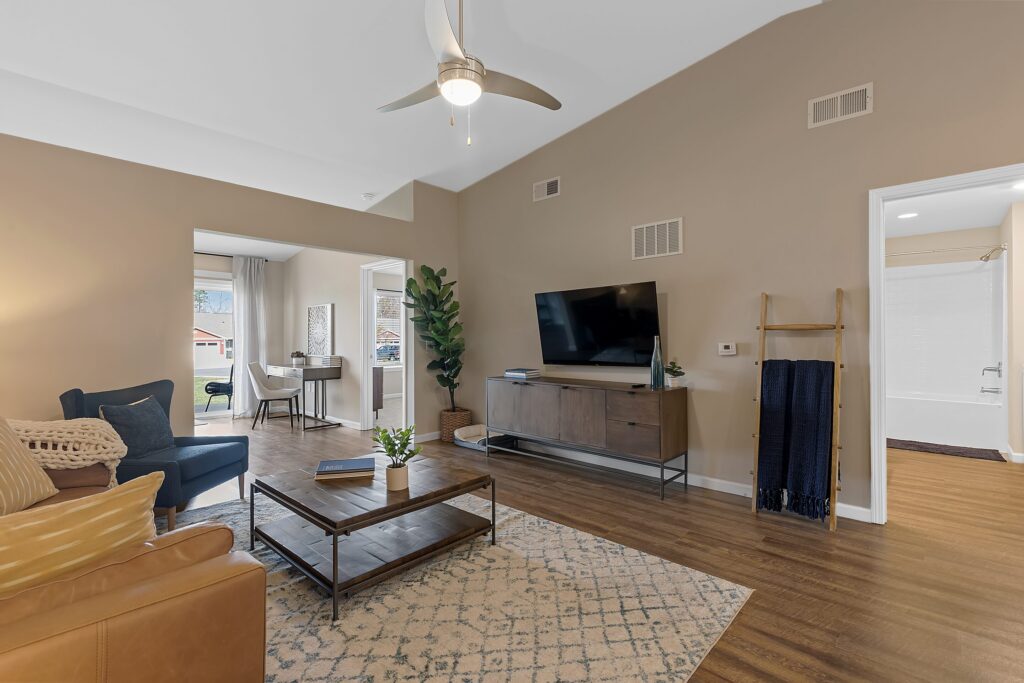 A modern living room with a ceiling fan, wall-mounted TV, coffee table, armchair, and sofa. An open doorway leads to a home office; a bathroom is visible through another door.