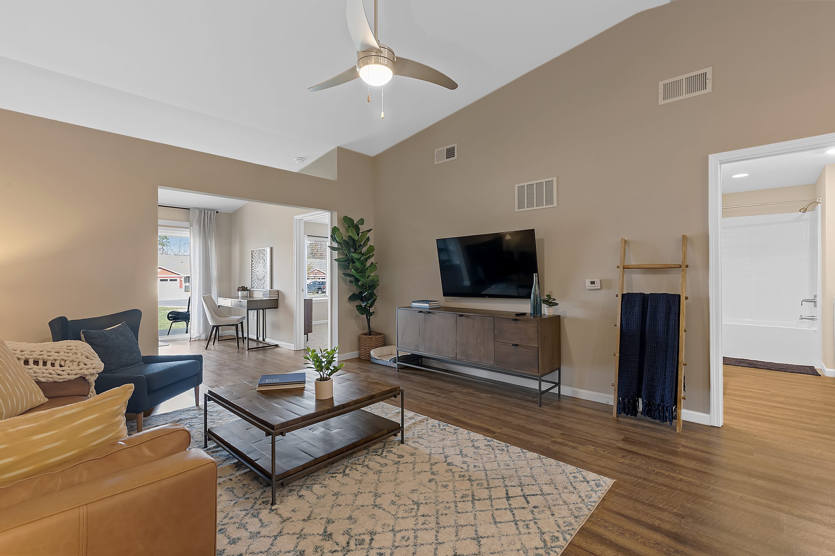 A modern living room with a ceiling fan, wall-mounted TV, coffee table, armchair, and sofa. An open doorway leads to a home office; a bathroom is visible through another door.