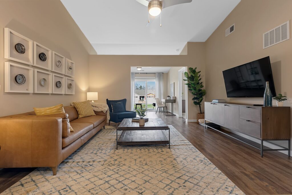 Modern living room with tan walls, brown leather sofa, patterned rug, wall-mounted TV, coffee table, and decorative wall art; dining area and window visible in the background.