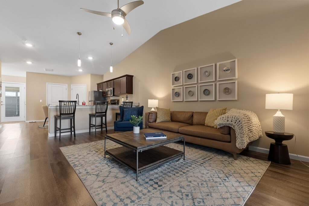 Modern open-plan living room and kitchen with wooden flooring, brown sofa, area rug, coffee table, bar stools, and pendant lights. Neutral tones and minimalistic decor.