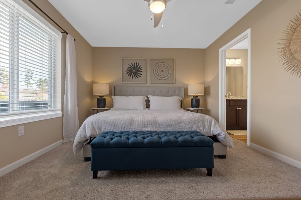A neatly made bed with white bedding, two bedside tables with lamps, a blue upholstered bench, and wall art in a beige-carpeted bedroom with natural light.