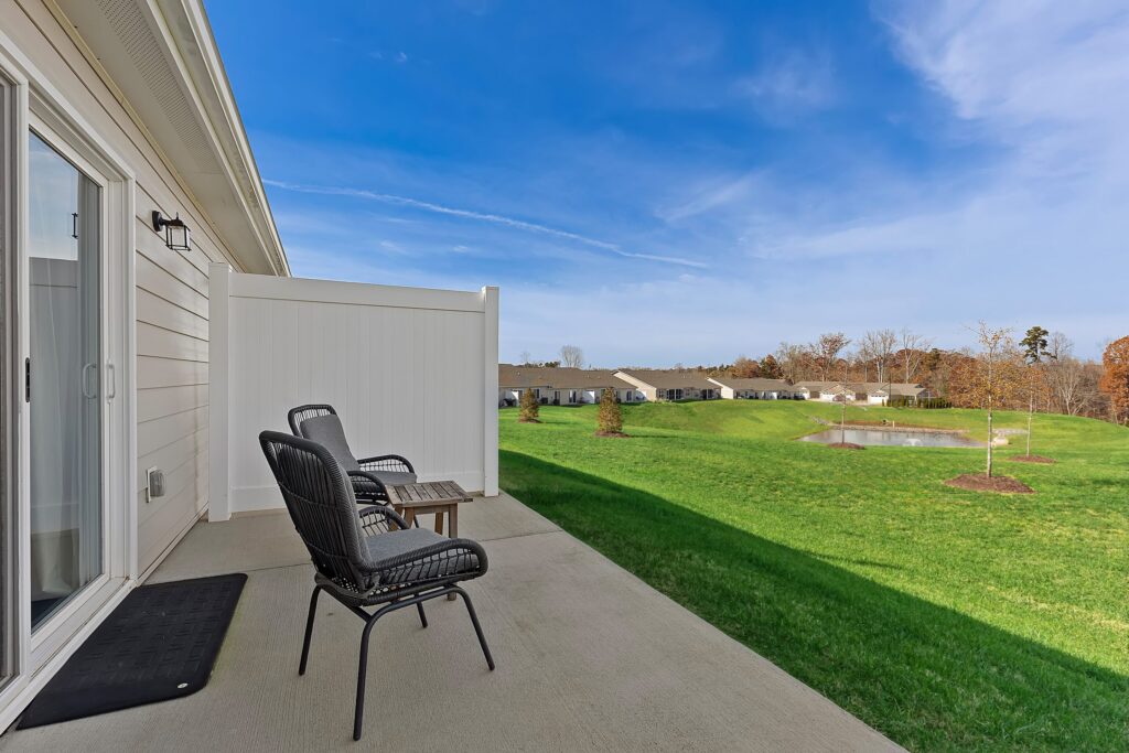 Two black patio chairs and a table on a concrete patio overlooking a grassy lawn, pond, and nearby row of houses under a blue sky.