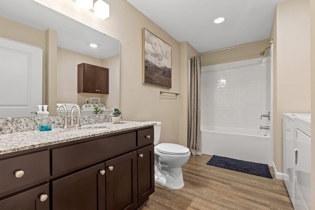 Modern bathroom with granite countertop, dark wood vanity, toilet, white bathtub with shower curtain, washing machine, and wood-look flooring. Neutral wall colors and a framed landscape painting.