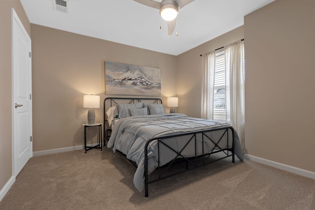 A bedroom with a metal bed frame, gray bedding, two bedside tables with lamps, beige walls, carpeted floor, a large window with sheer curtains, and abstract wall art.